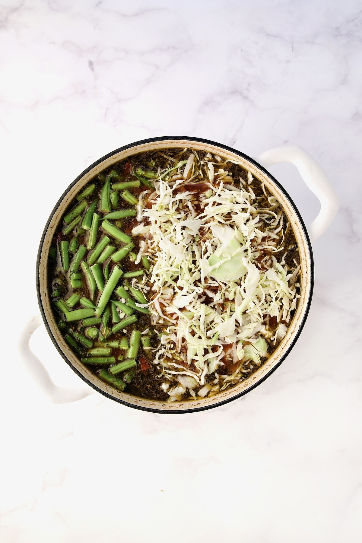 Green beans and cabbage added to soup in process in white Dutch oven.