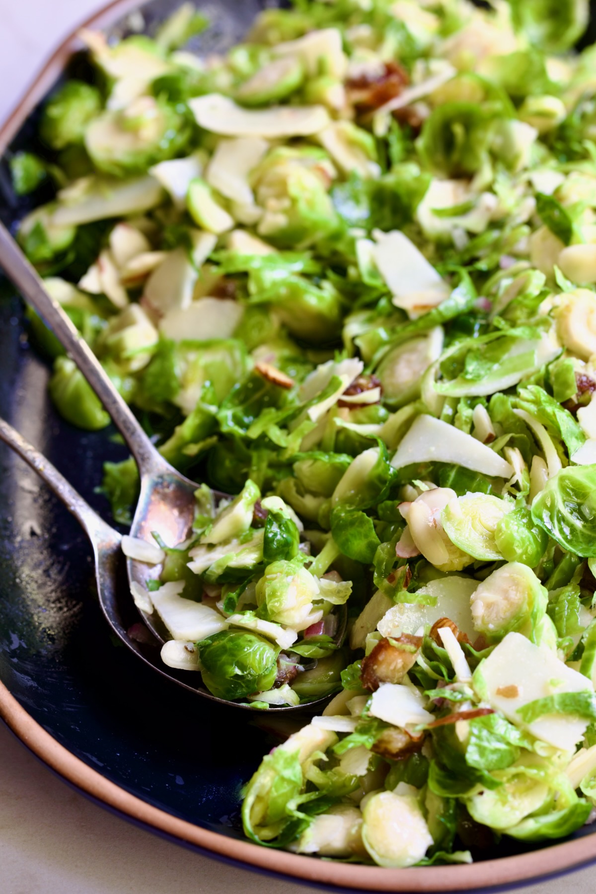 Shaved Brussels Sprouts Salad on a black platter with brown rim being scooped up with serving utensils.