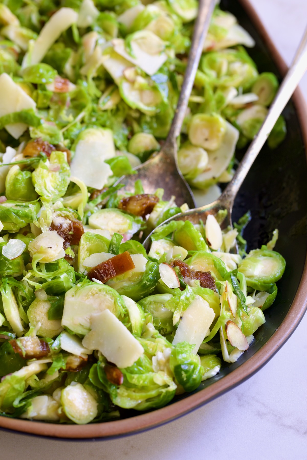 Shaved Brussels Sprouts Salad on a black platter with brown rim being scooped up with serving utensils.