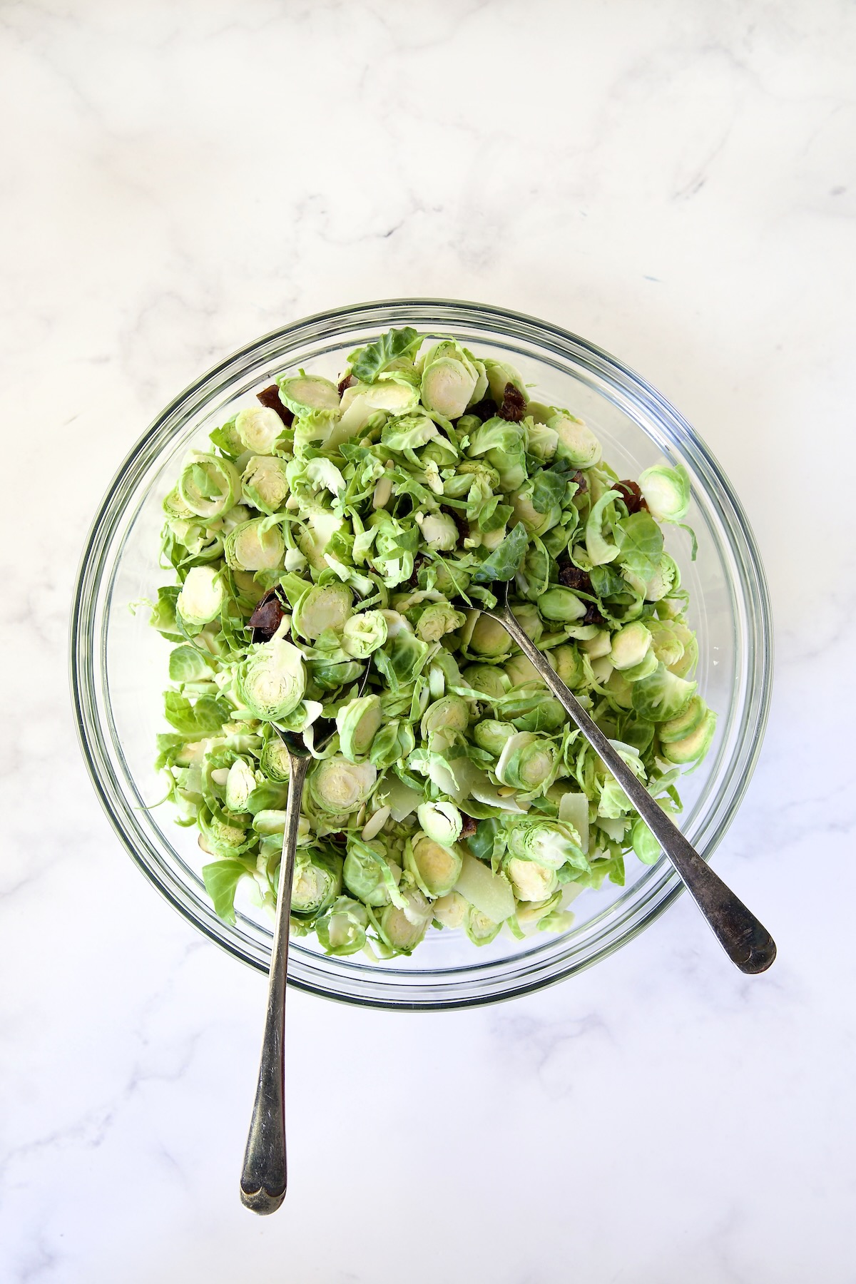 Shaved Brussels Sprouts Salad in glass bowl before dressing is added being mixed together with serving utensils.