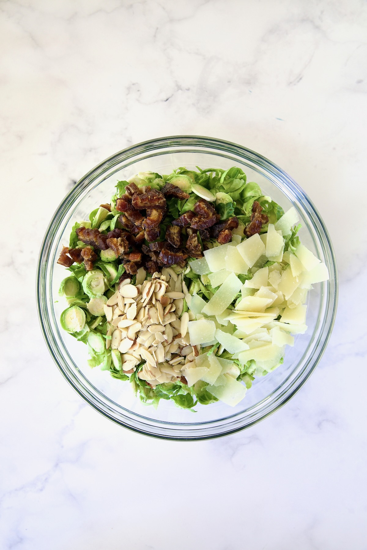 Shaved Brussels sprouts, chopped dates, sliced almonds and shaved Romano cheese in glass bowl before being mixed together.