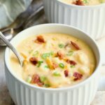 Cheddar Bacon Potato Soup in large white soup mug with spoon garnished with bacon, scallions and cheese shreds.
