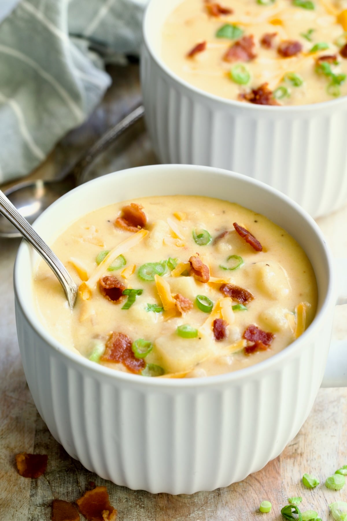 Cheddar Bacon Potato Soup in large white soup mug with spoon garnished with bacon, scallions and cheese shreds.