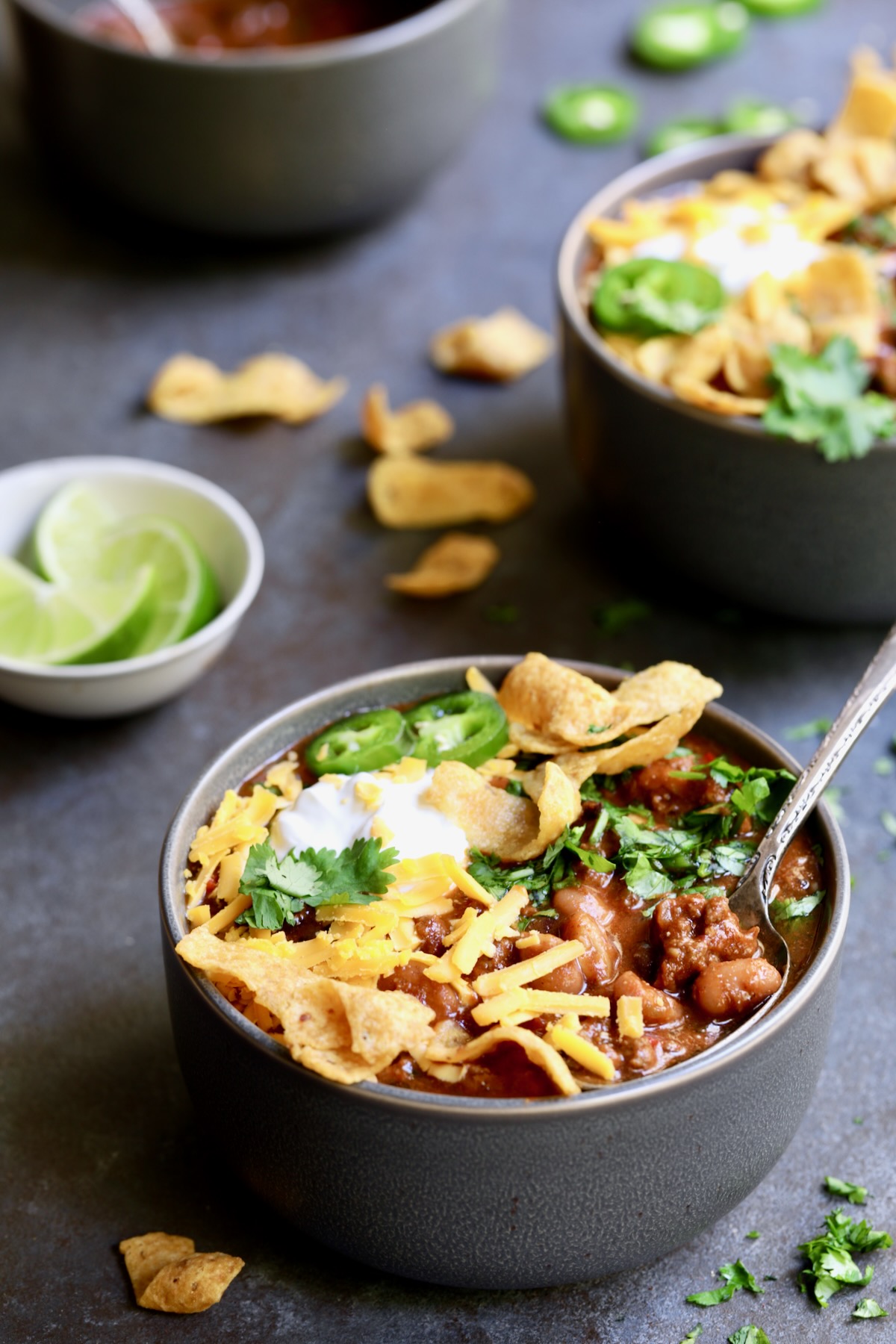 Bowls of Game Day Chili with corn chips, cheese, sour cream jalapeno slices and cilantro in gray bowl.