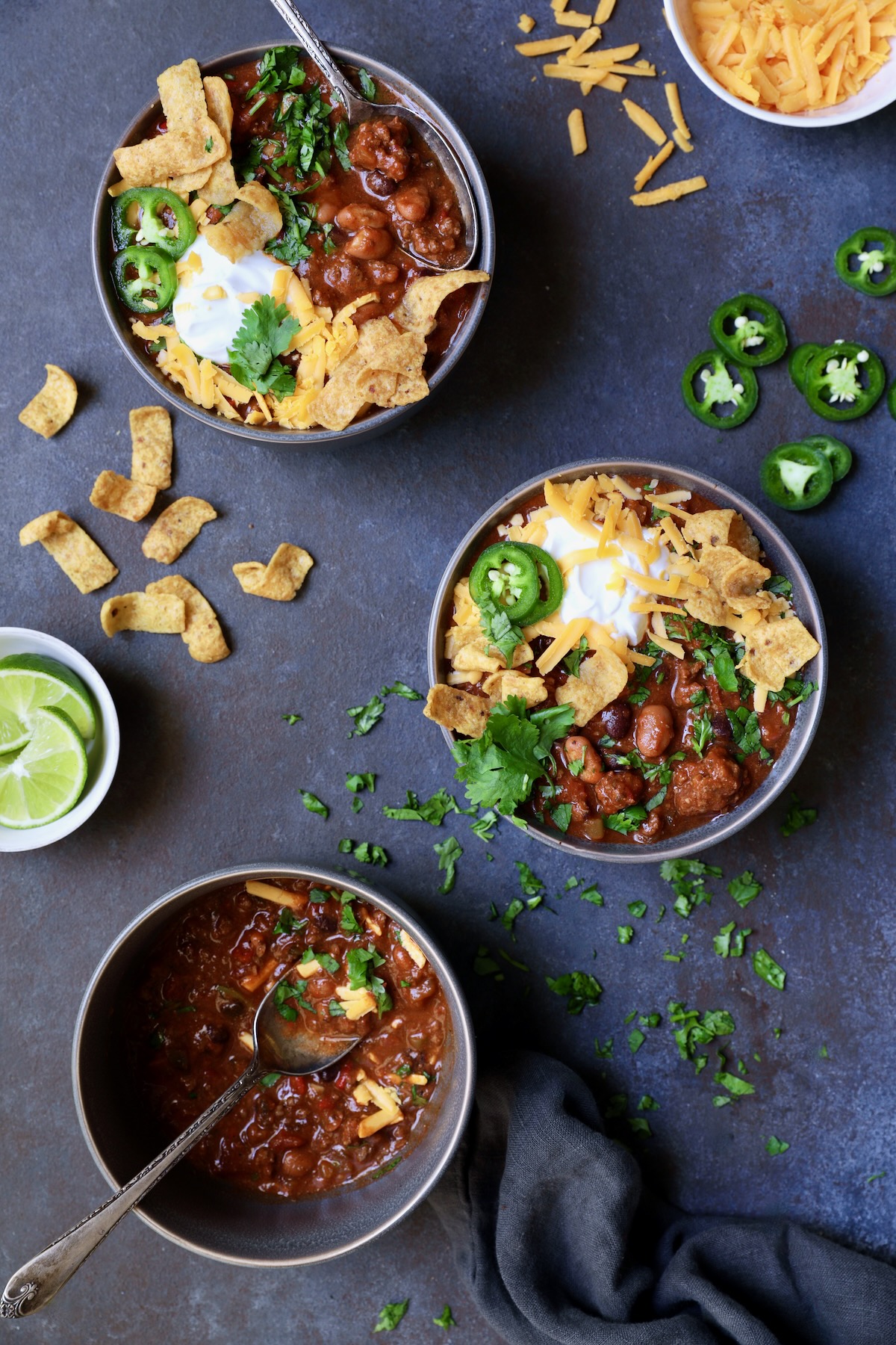 Three bowls of Game Day Chili with corn chips, cheese, sour cream jalapeno slices and cilantro in gray bowl.