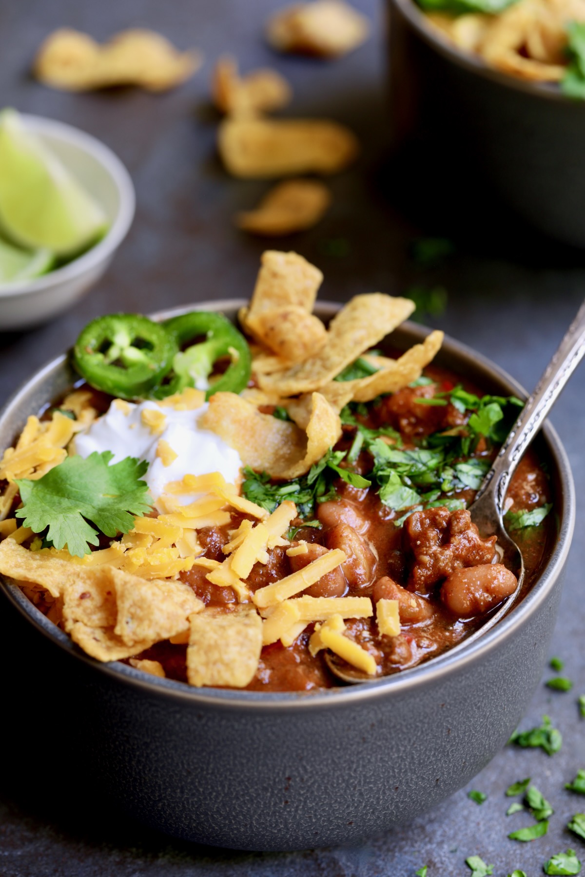 One bowl of Game Day Chili with corn chips, cheese, sour cream jalapeno slices and cilantro in gray bowl.