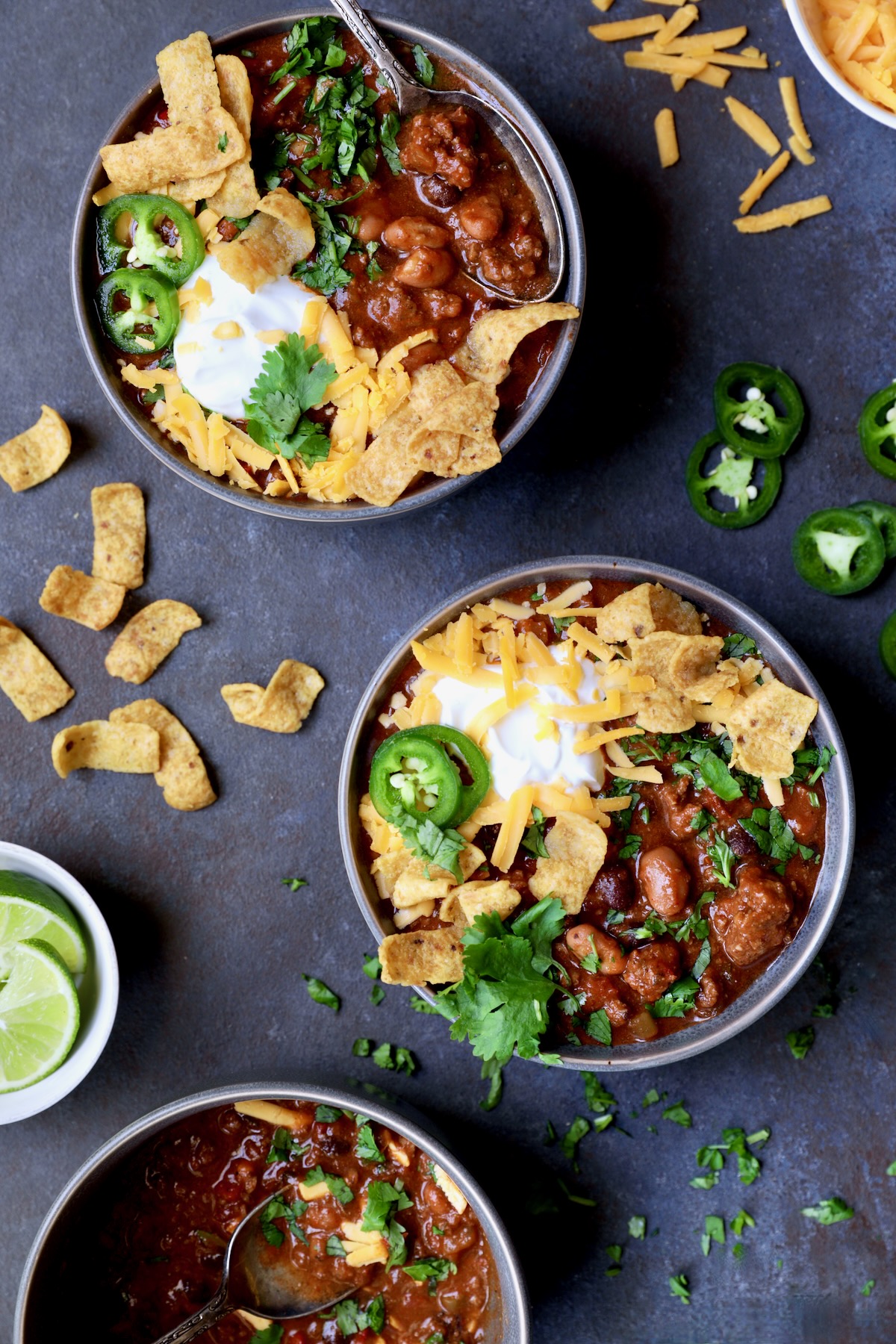 Two bowls of Game Day Chili with corn chips, cheese, sour cream jalapeno slices and cilantro in gray bowl.