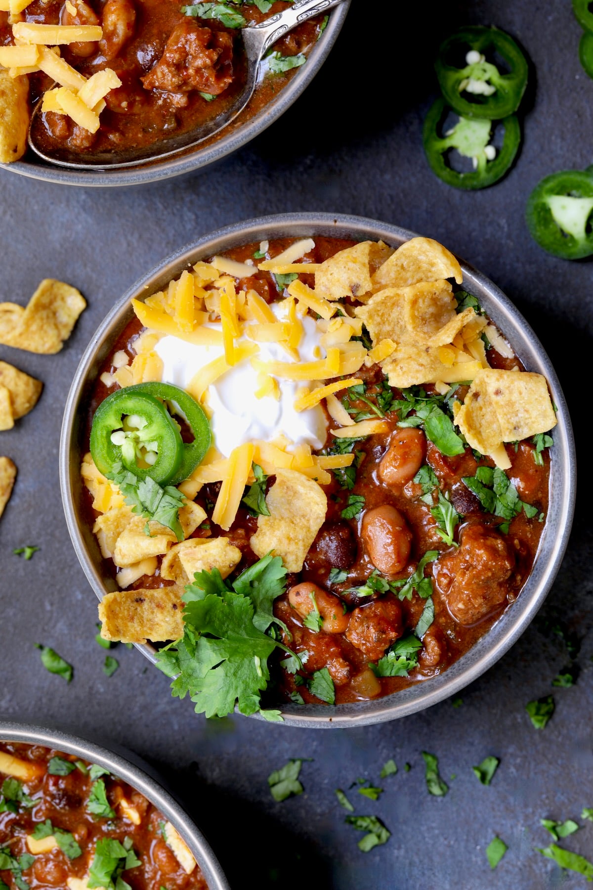 One bowl of Game Day Chili with corn chips, cheese, sour cream jalapeno slices and cilantro in gray bowl.