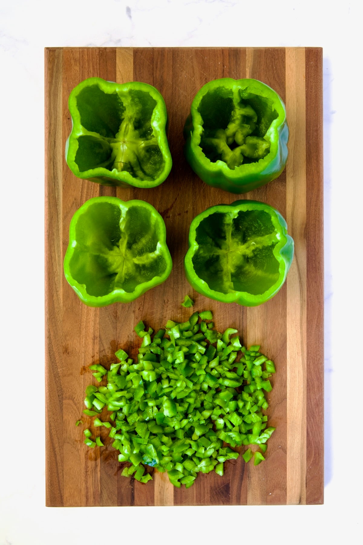 Four uncooked green bell peppers with the tops cut off and chopped green pepper on wood cutting board.
