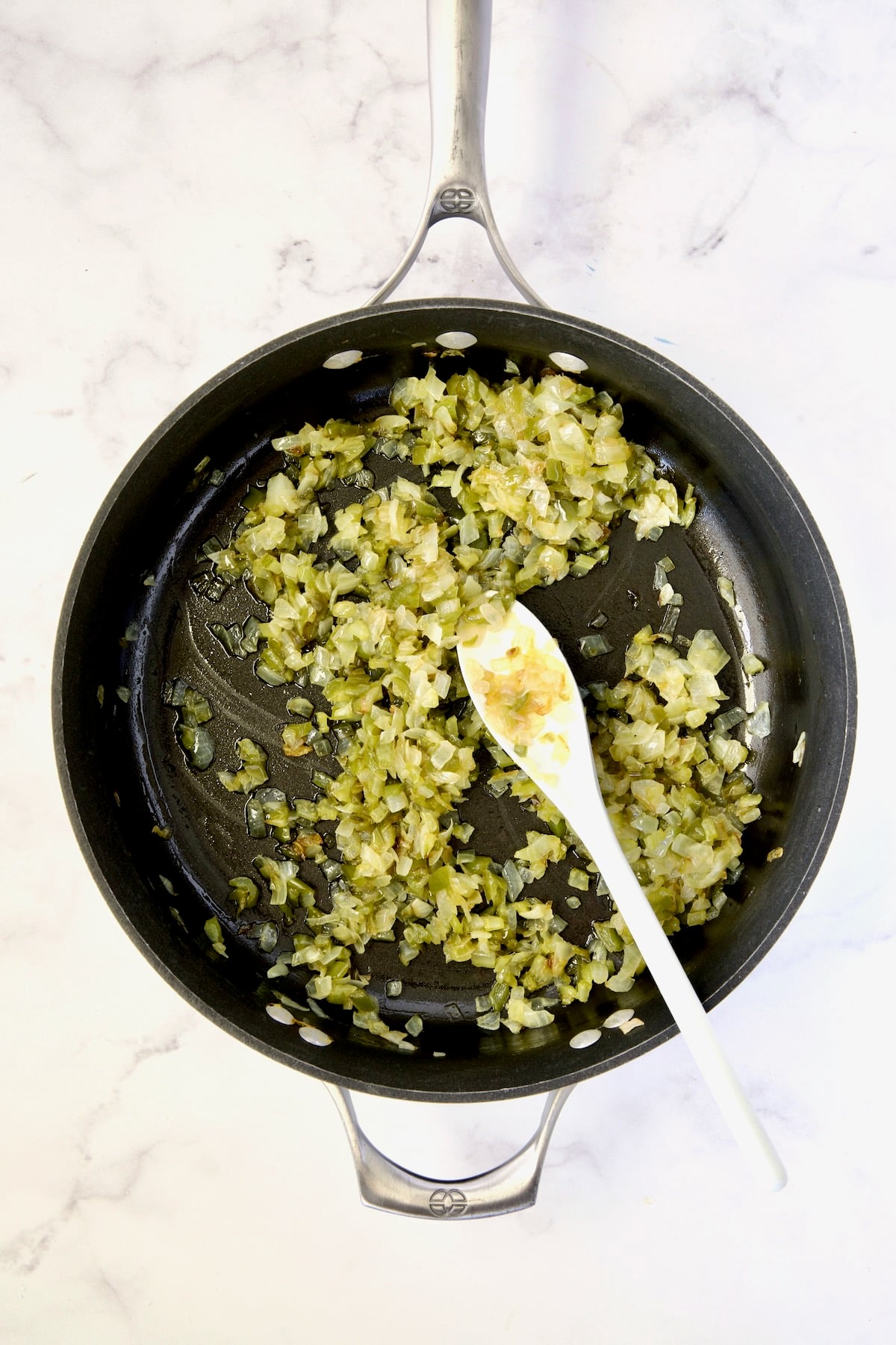 Cooked onions, bell pepper and celery in nonstick skillet being stirred with white spoon.