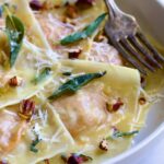 Pumpkin Ravioli with Sage Brown Butter in white pasta bowl garnished with toasted pecans and fried sage leaves.