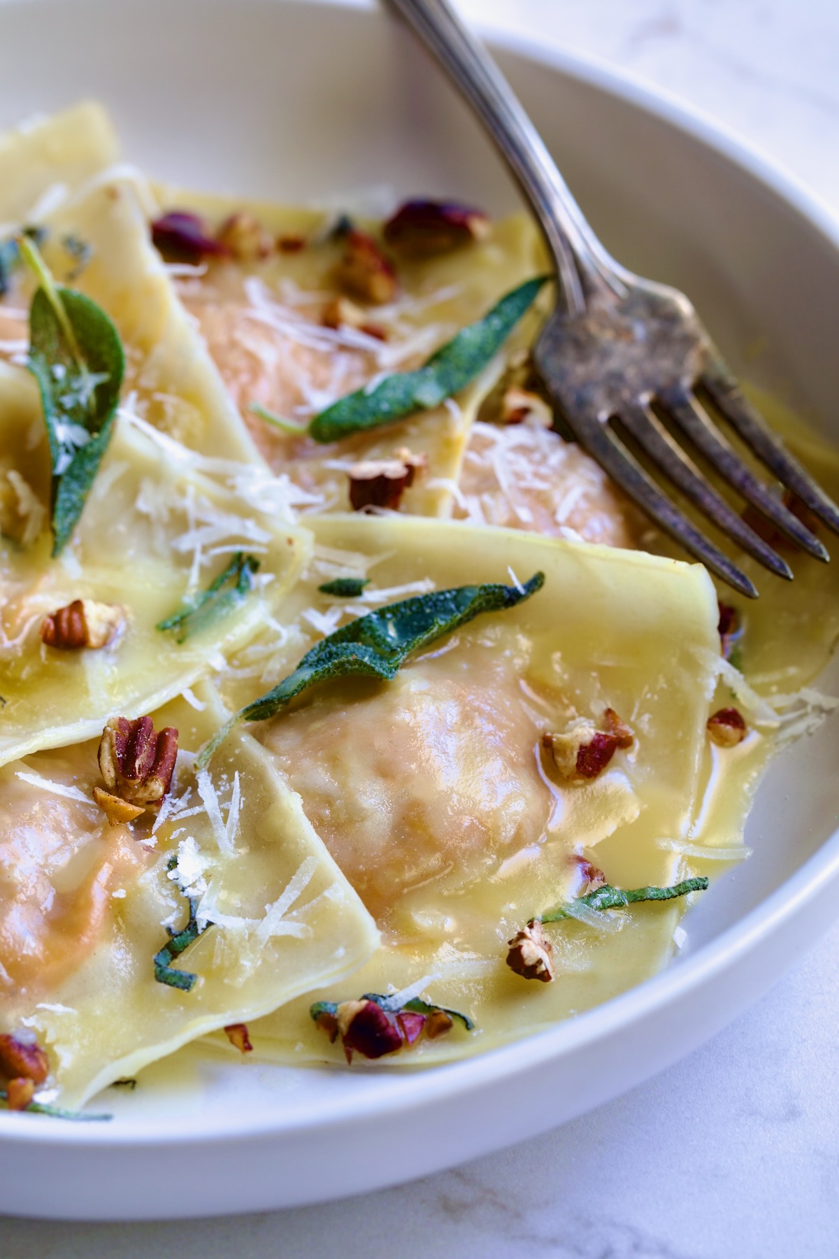 Pumpkin Ravioli with Sage Brown Butter in white pasta bowl garnished with toasted pecans and fried sage leaves.