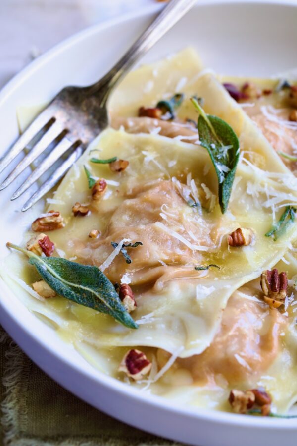 Pumpkin Ravioli with Sage Brown Butter in white pasta bowl garnished with toasted pecans and fried sage leaves.