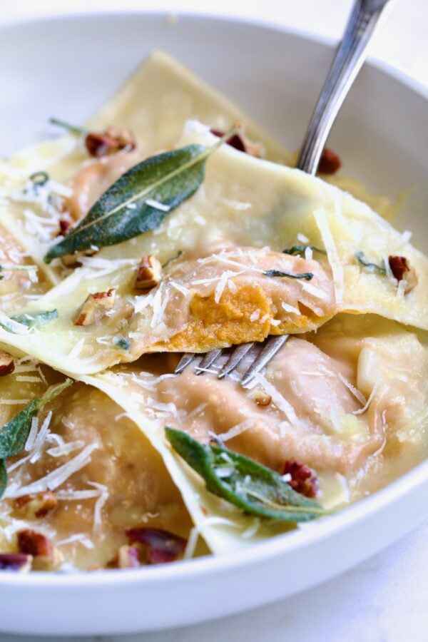 Pumpkin Ravioli with Sage Brown Butter in white pasta bowl garnished with toasted pecans and fried sage leaves with one of the raviolis cut into to expose the filling.