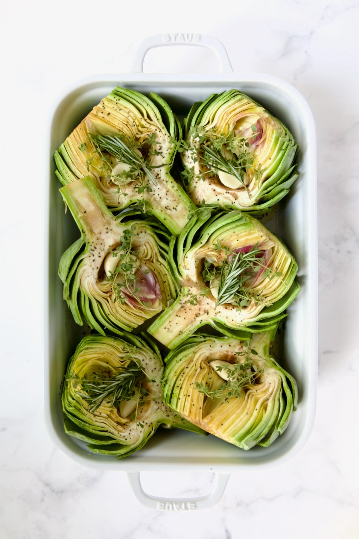 Six artichoke halves in a white roasting pan with garlic and herbs on top of them.