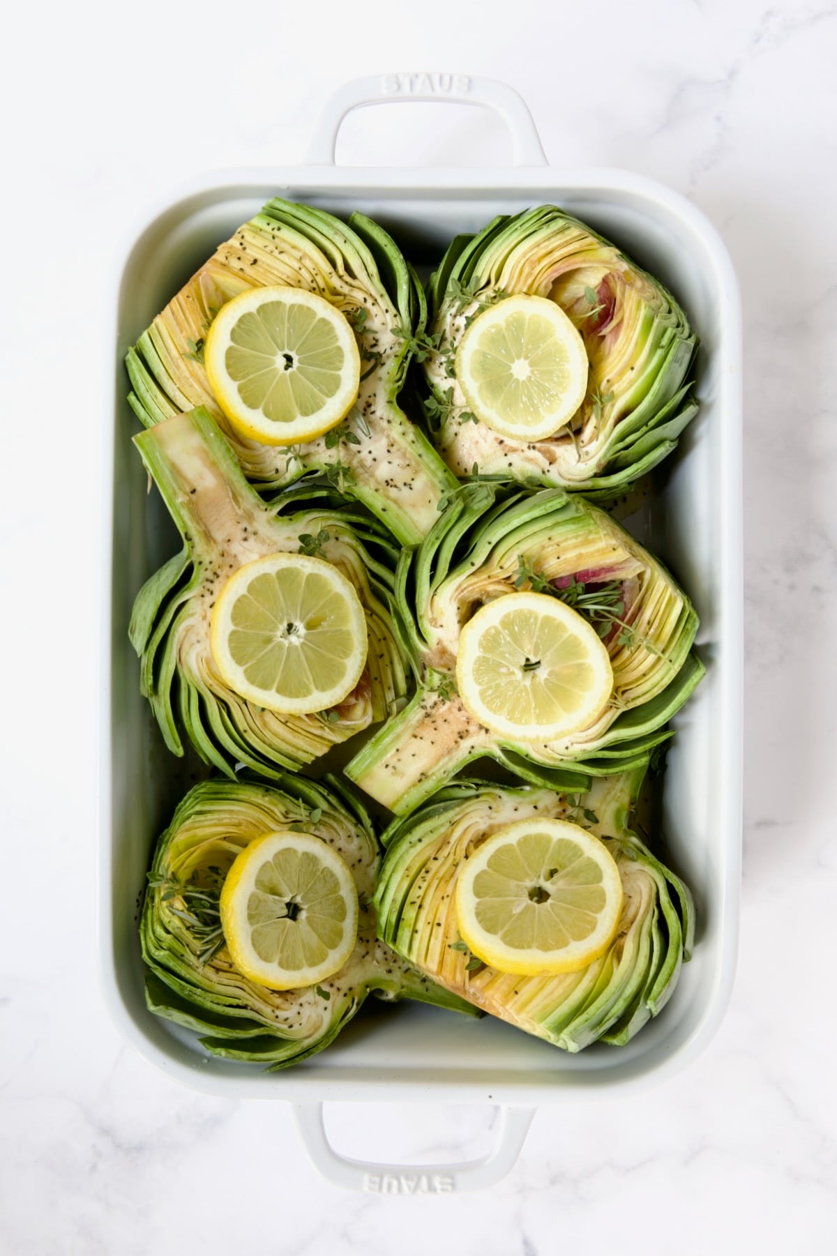 Six artichoke halves in a white roasting pan with lemon slices set atop the herbs and garlic cloves.