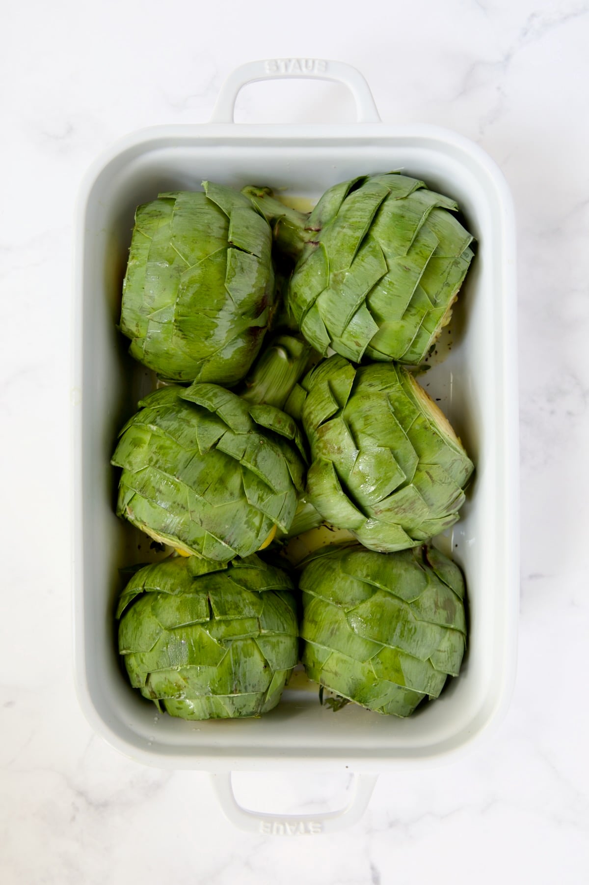 Six artichoke halves sitting cut side down in a white roasting pan.