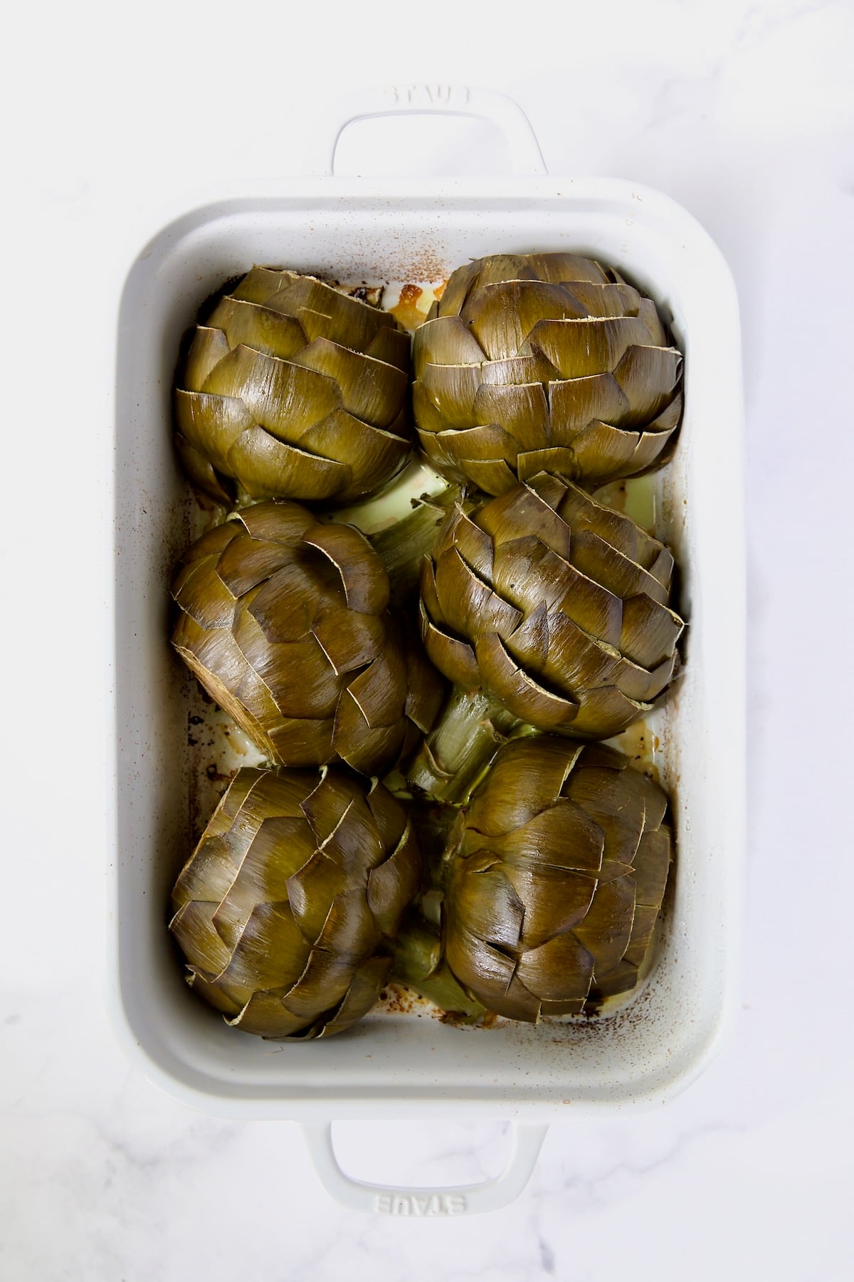 Six artichoke halves in a white roasting pan with cut side down after being roasted.