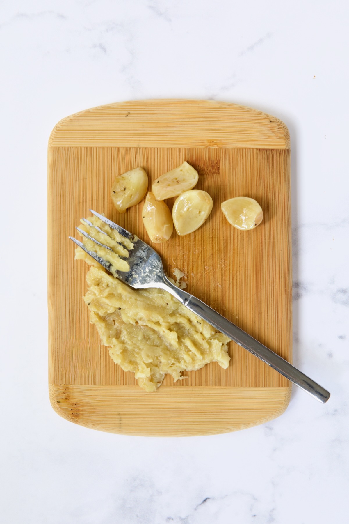 Garlic being smashed on bamboo cutting board with fork.