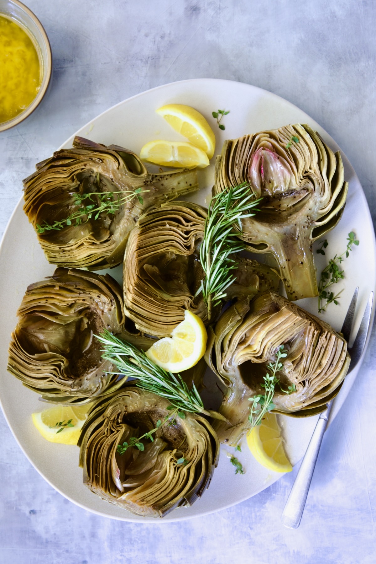 Six Roasted Artichoke halves with Garlic Butter garnished with rosemary, thyme sprigs and lemon wedges.