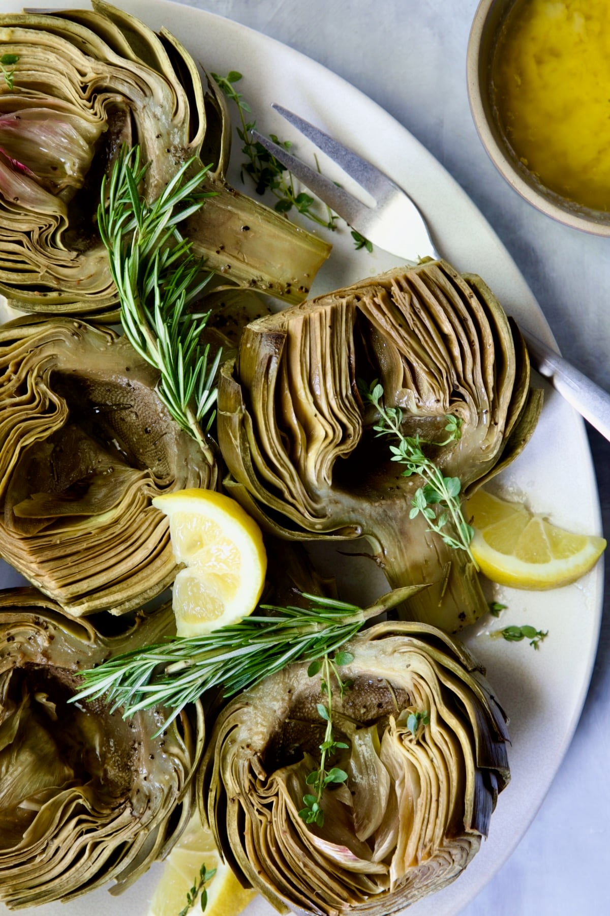 Roasted Artichokes with Garlic Butter on white oval platter garnished with herb sprigs and lemon wedges.