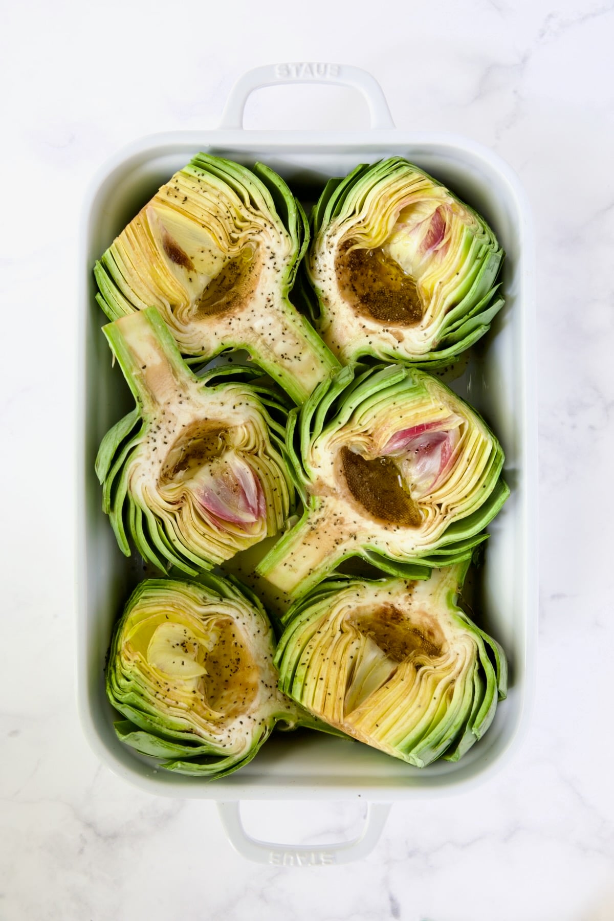 Six artichoke halves in a white roasting pan drizzled with olive oil.