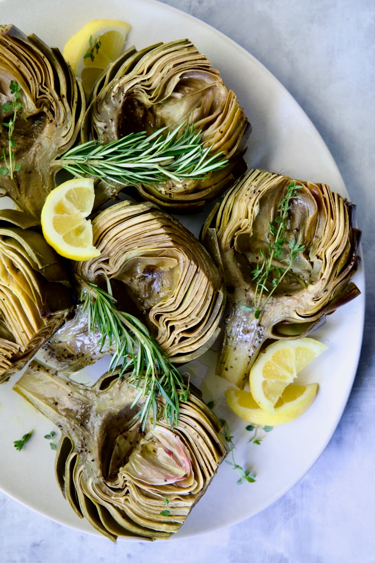 Roasted Artichokes with Garlic Butter on white oval platter garnished with lemon wedges and herb sprigs.