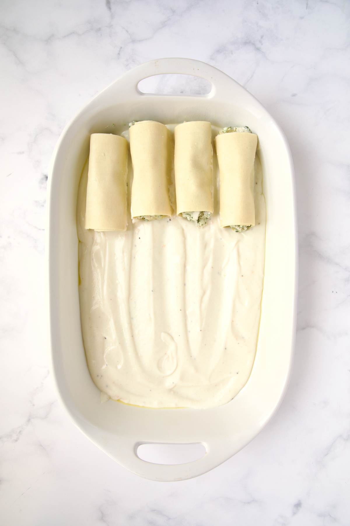 Four cannelloni set on bechamel sauce in white oval baking dish.