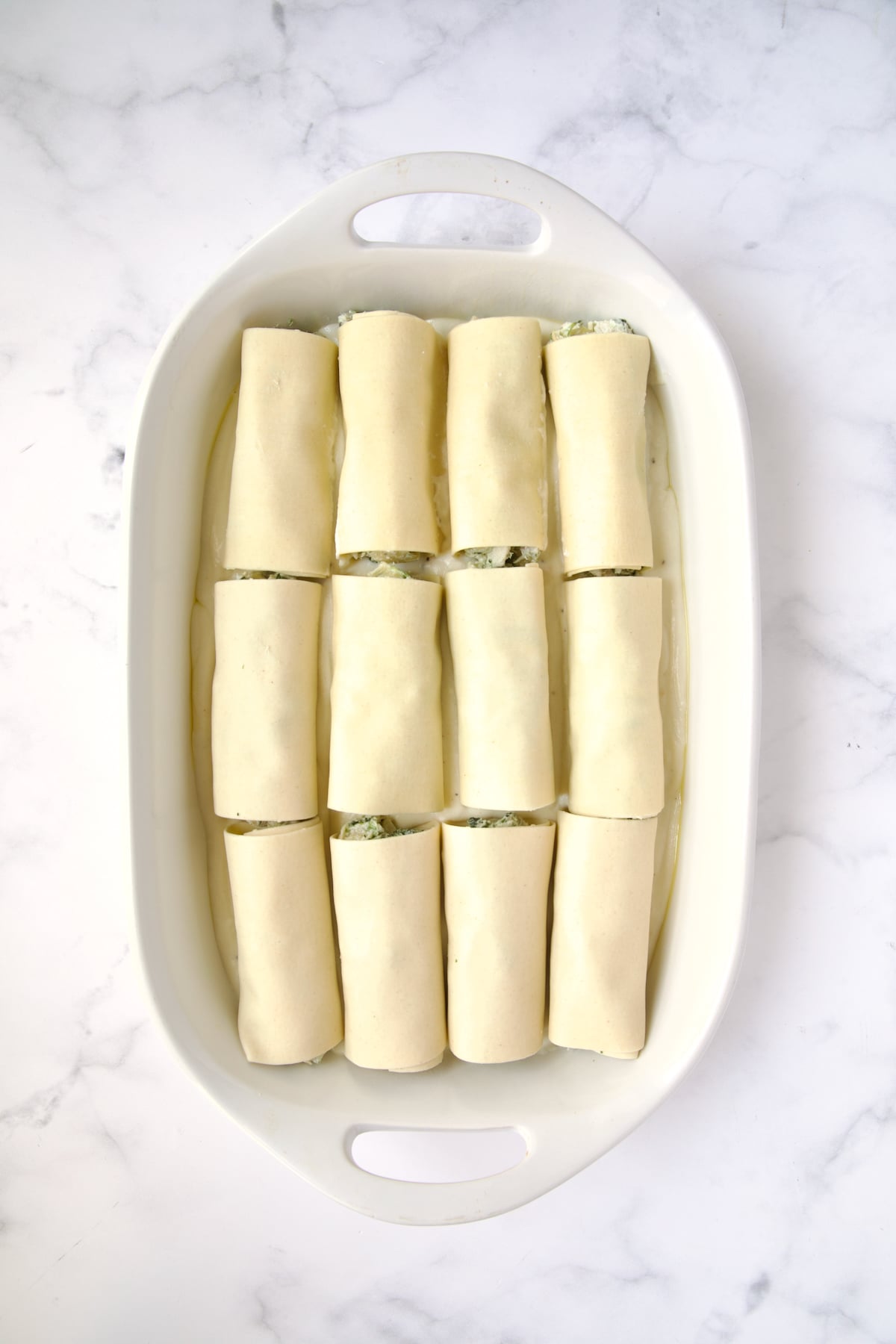 12 cannelloni set on bechamel sauce in white oval baking dish.