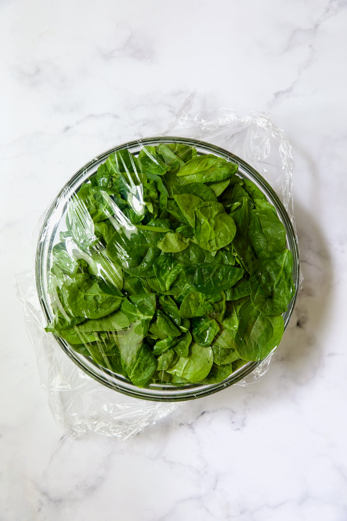 Glass bowl full of spinach with plastic wrap over the top.