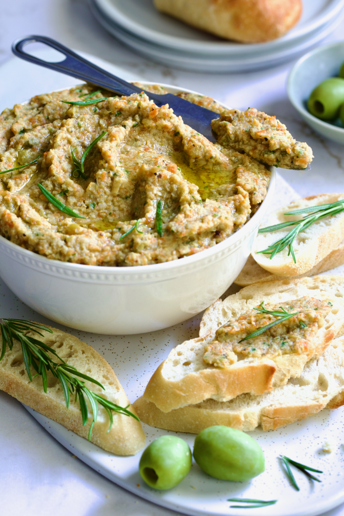 Green Olive Eggplant Tapenade in white bowl with serving knife and bread slices scattered about.