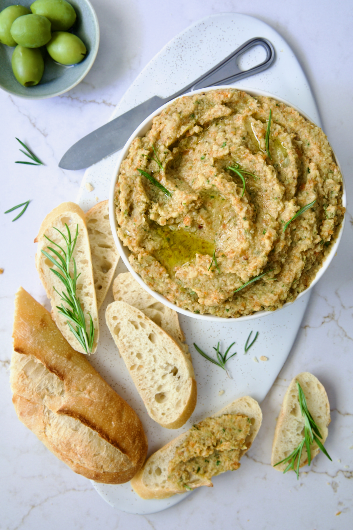 Green Olive Eggplant Tapenade in white bowl with serving knife and bread slices scattered about garnished with fresh rosemary.