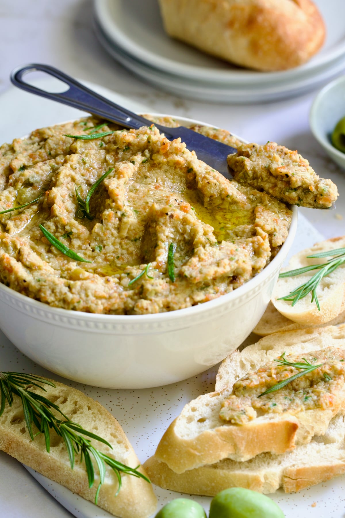 Green Olive Eggplant Tapenade in white bowl with serving knife and bread slices scattered about garnished with fresh rosemary.