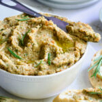 Green Olive Eggplant Tapenade in white bowl with serving knife and bread slices scattered about.