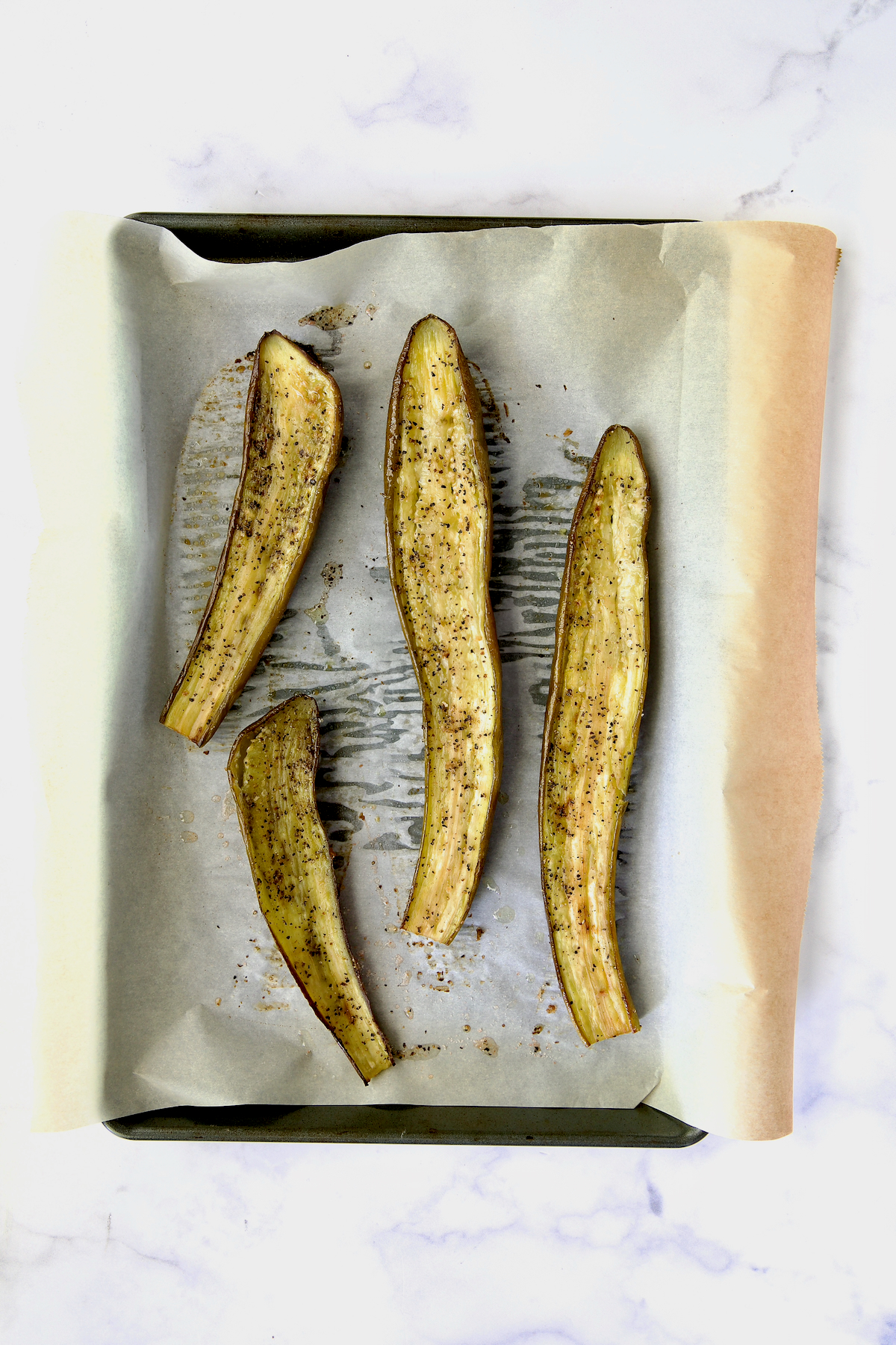 Four slices of Japanese eggplant cut side up after roasting on parchment paper-lined rimmed baking sheet.