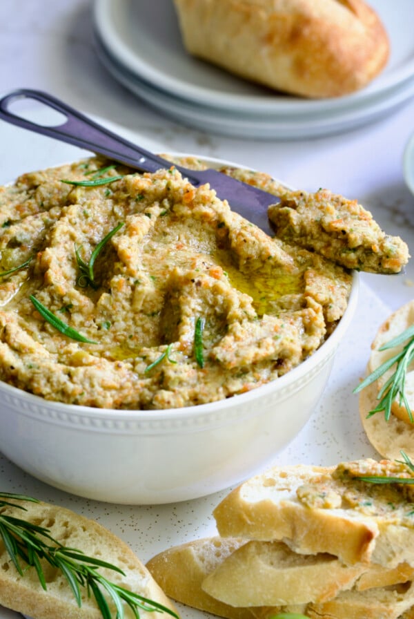 Green Olive Eggplant Tapenade in white bowl with serving knife and bread slices scattered about.