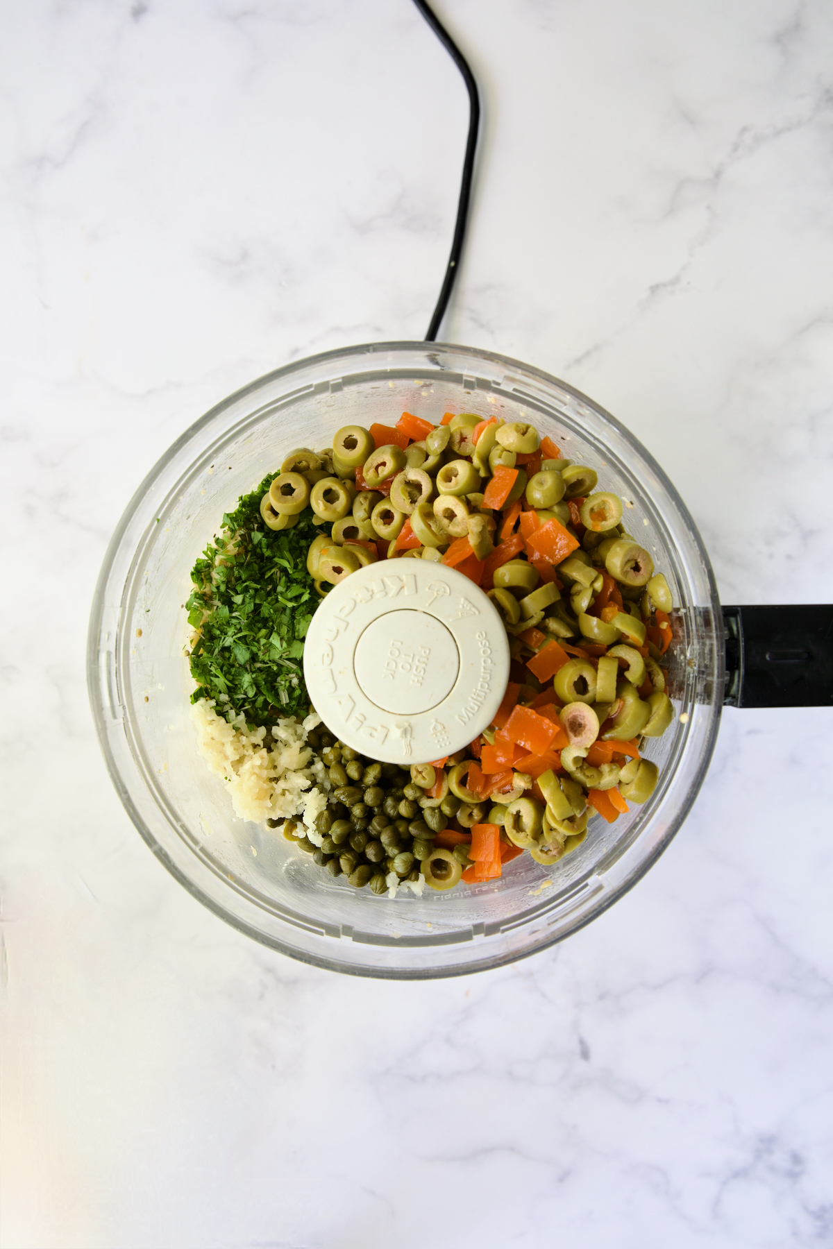 Olives, garlic, capers, parsley, and rosemary added to bowl of food processor with pureed eggplant and chickpeas before being fully pureed.