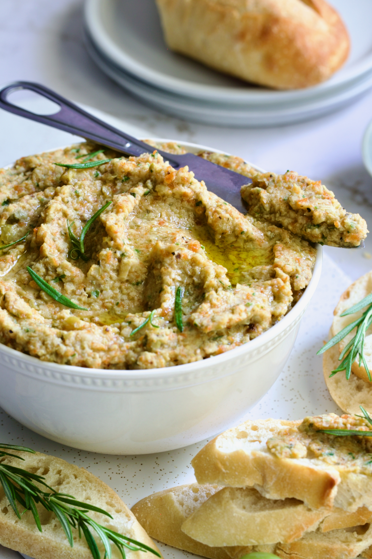 Green Olive Eggplant Tapenade in white bowl with serving knife and bread slices scattered about.