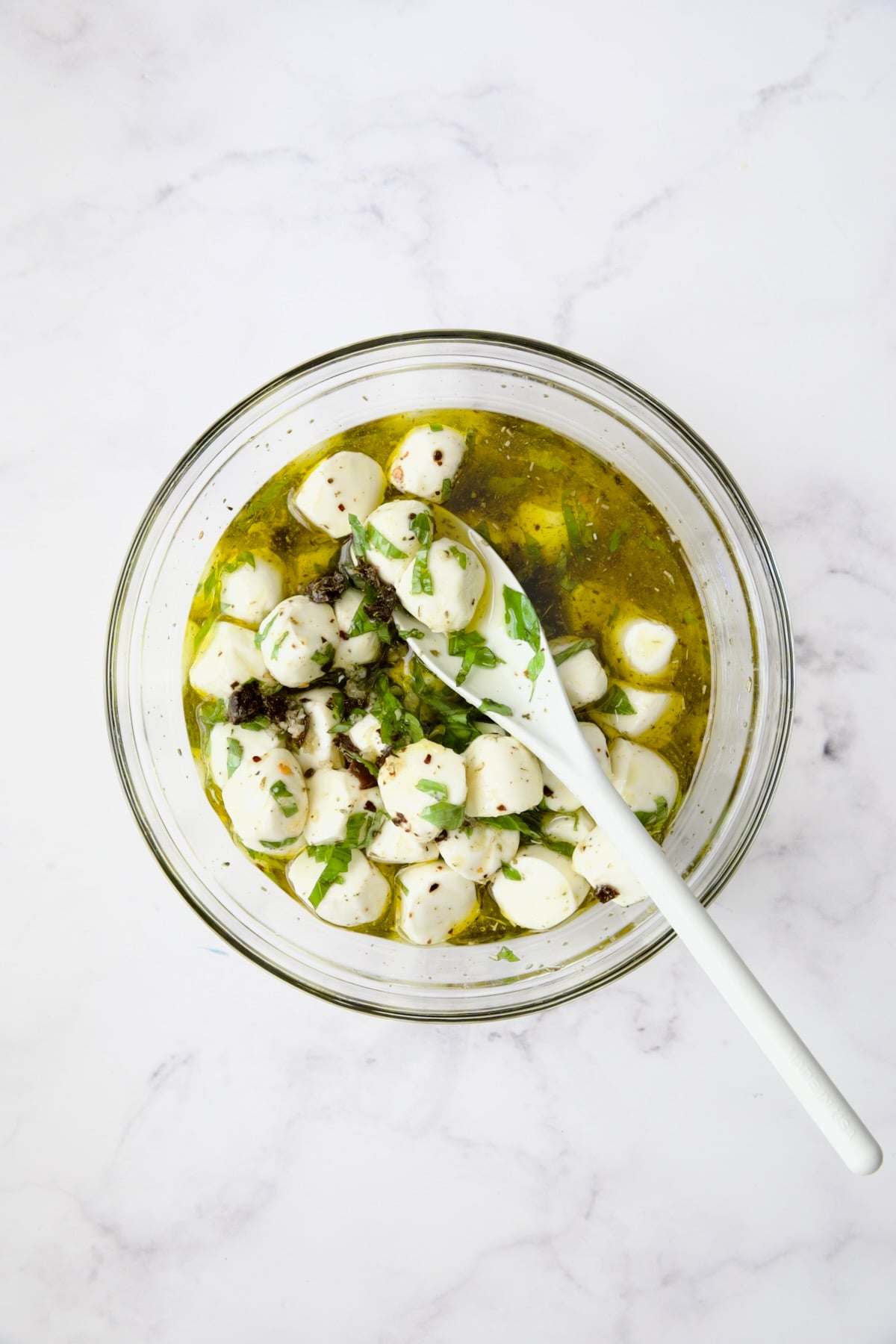 Fresh mozzarella balls and marinade being stirred with white spoon in glass bowl.