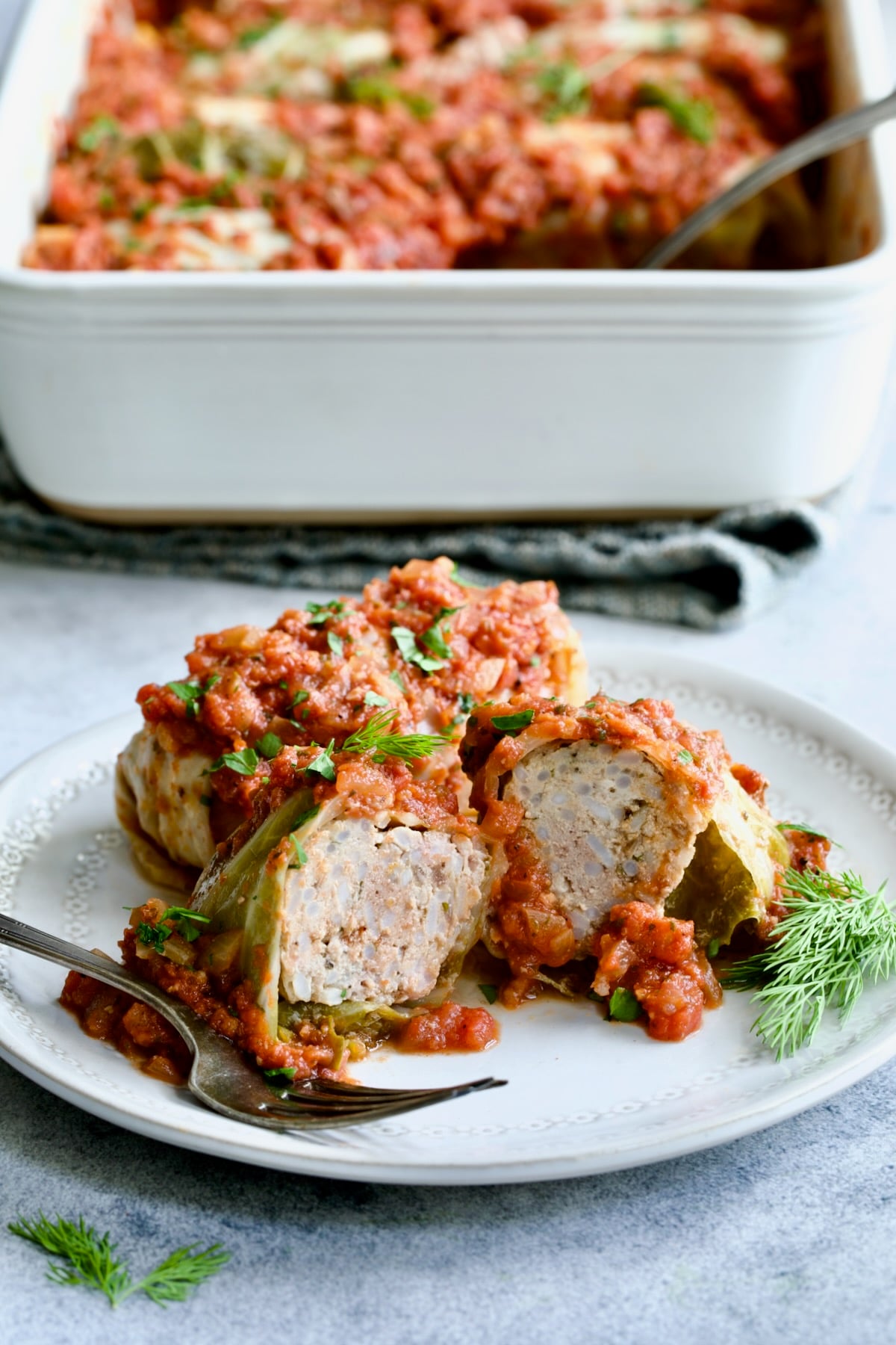 Polish Stuffed Cabbage roll cut in half to expose filling on white plate with baking dish of cabbage in the background.