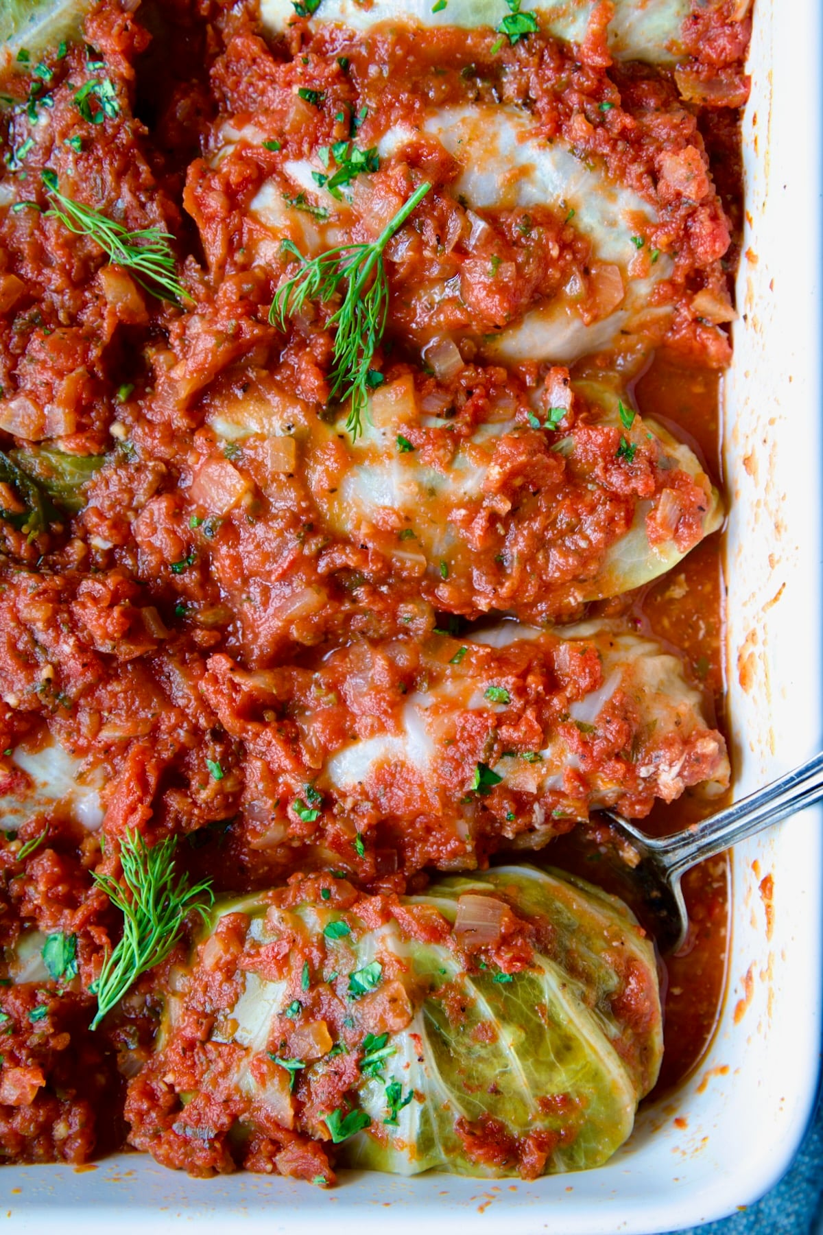 Row of four Polish Stuffed Cabbage in white baking dish with serving fork garnished with fresh dill.