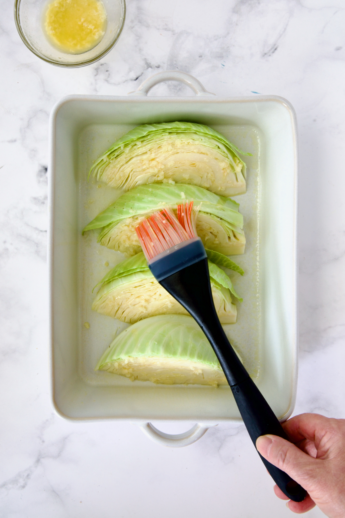 Four wedges of cabbage in white baking dish being brushed with butter and garlic.