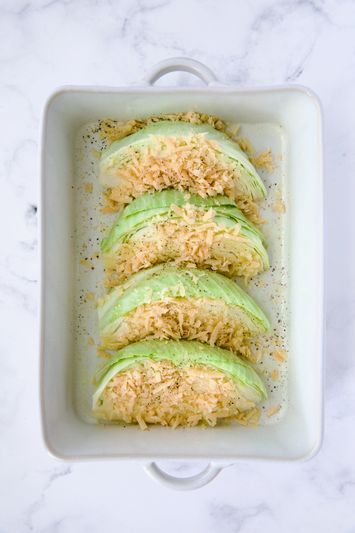 Four wedges of cabbage in white rectangular baking dish sprinkled with Parmesan cheese.