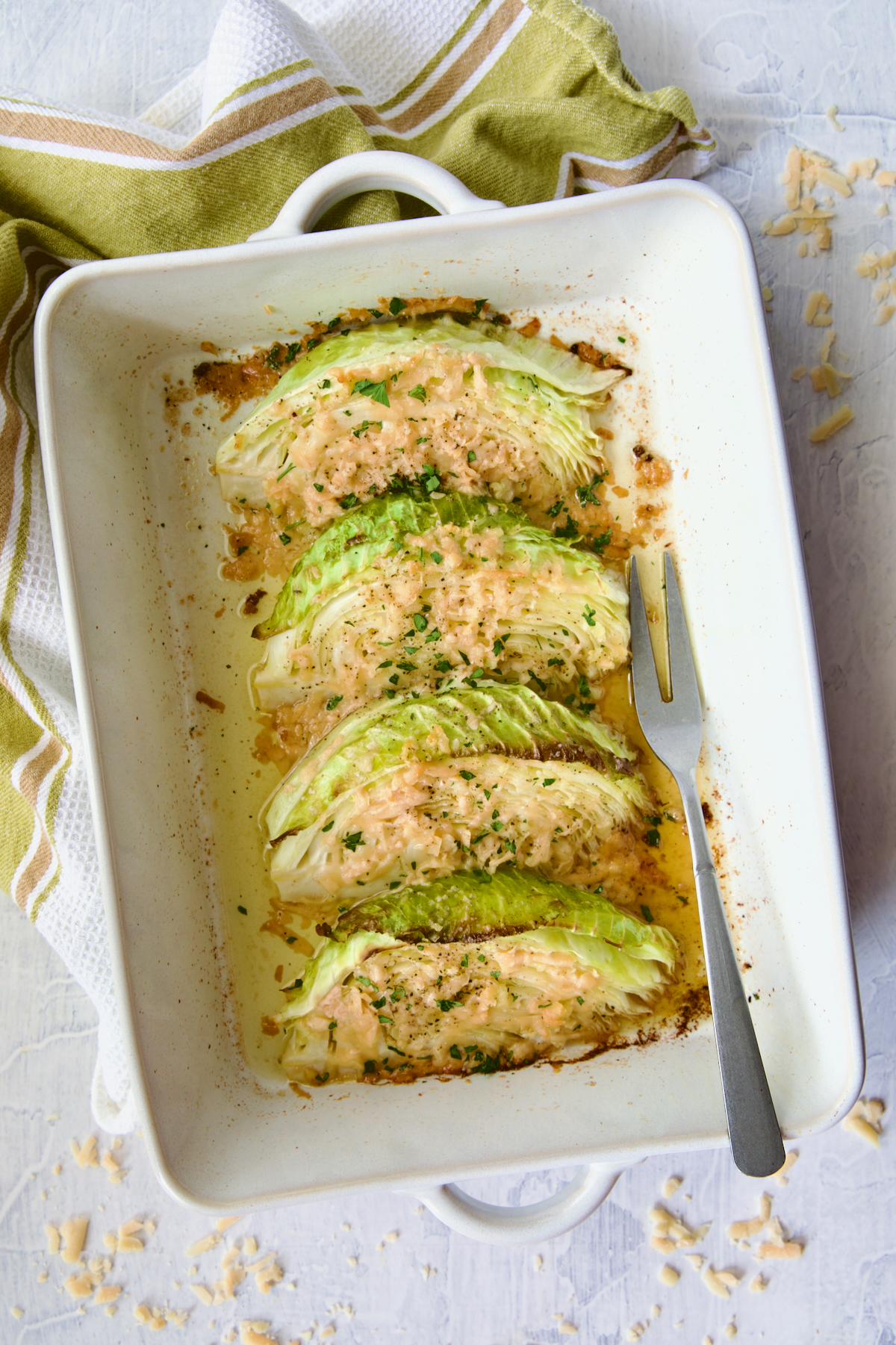 Baked Garlic Parmesan Cabbage Wedges in white rectangular baking dish with serving fork and green striped towel.