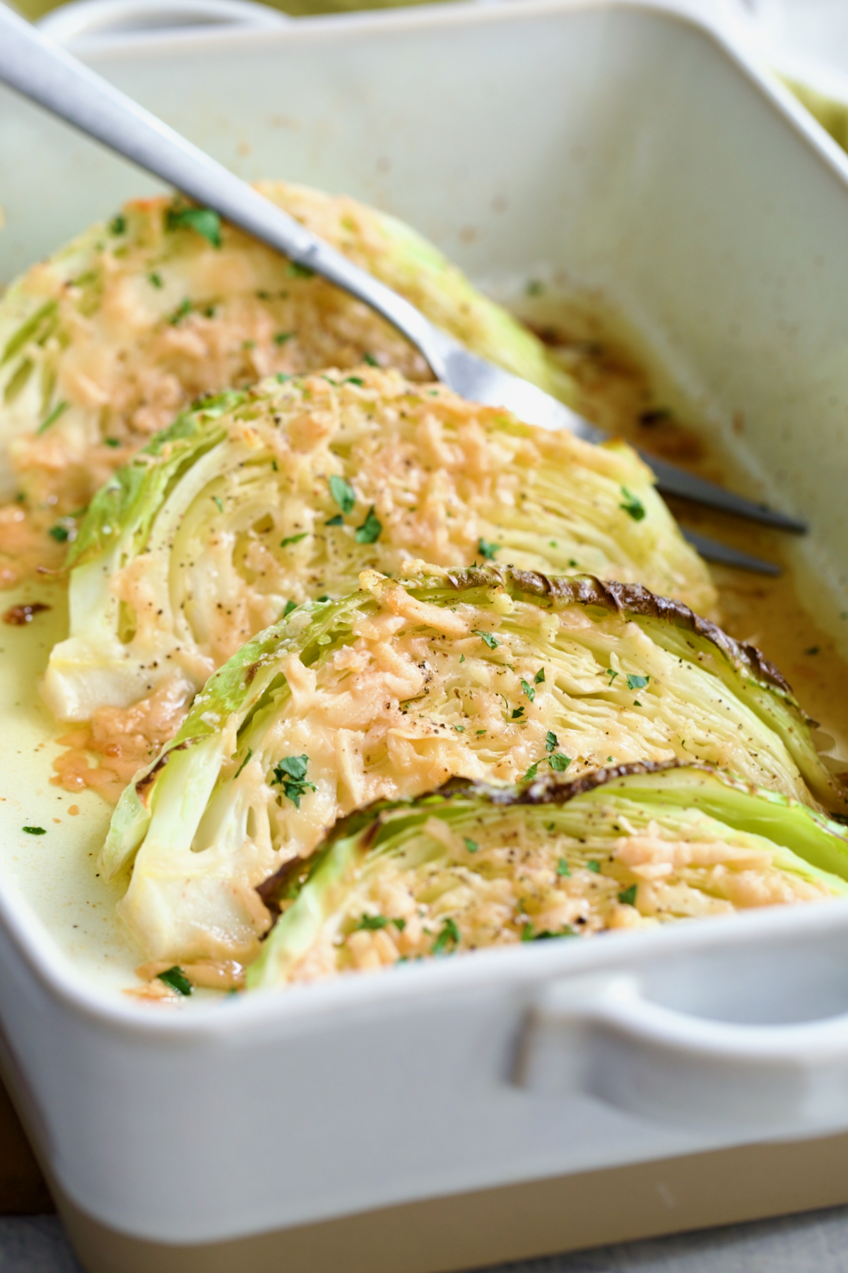 Four wedges of Baked Garlic Parmesan Cabbage Wedges in baking dish with serving fork garnished with fresh chopped parsley.
