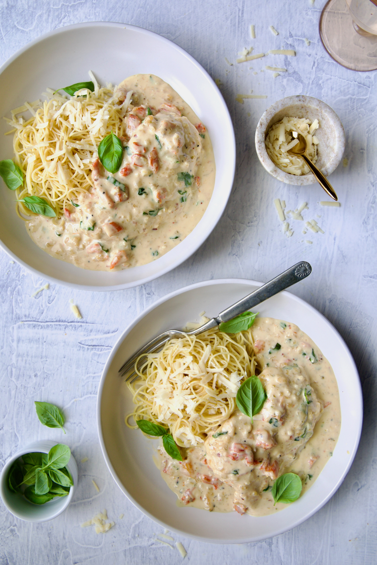 Two servings of Marry Me Chicken in white bowls with angel hair pasta and fork garnished with fresh basil leaves.