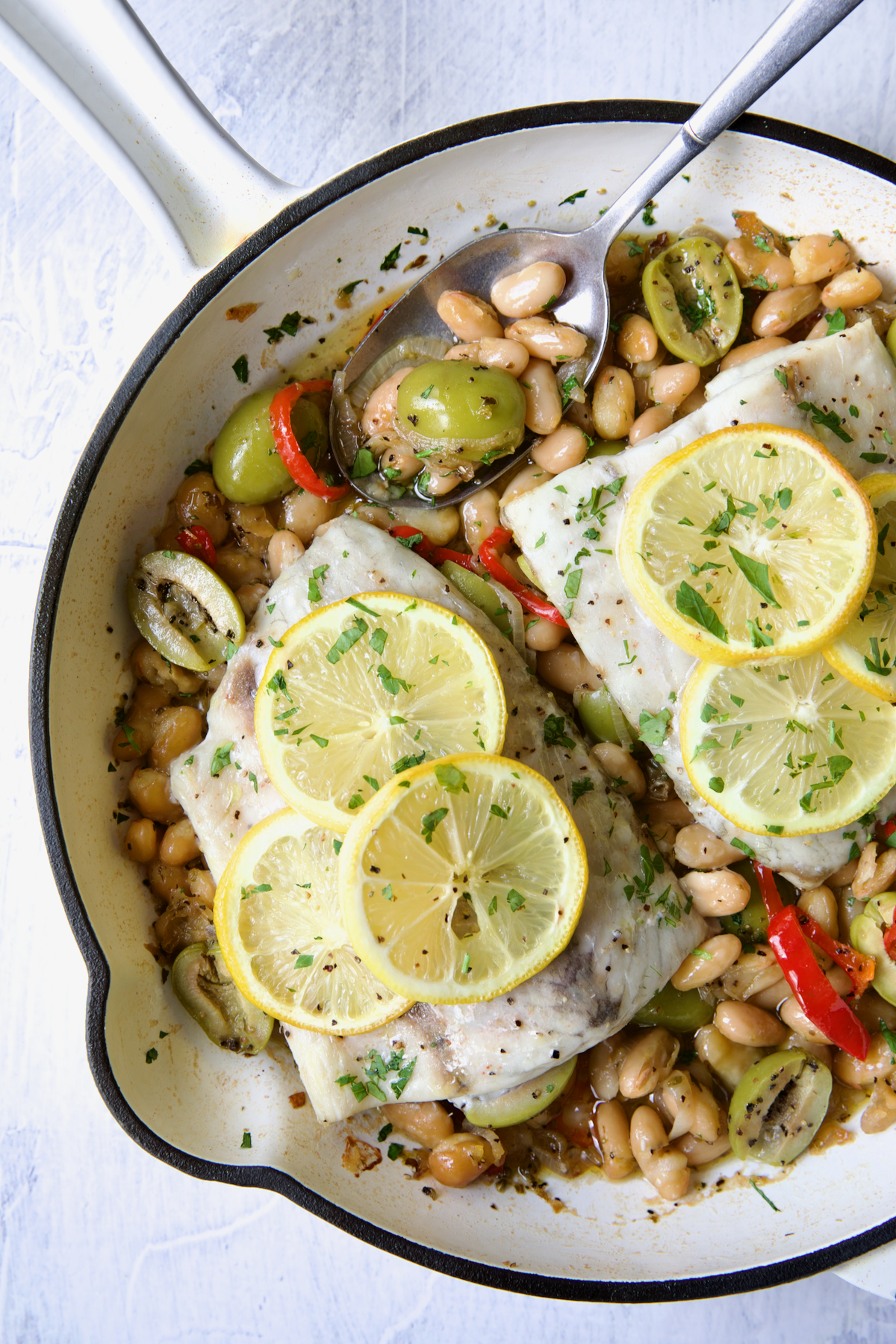 Mediterranean Fish and White Bean Skillet in white cast iron skillet topped with lemon slices with serving spoon.
