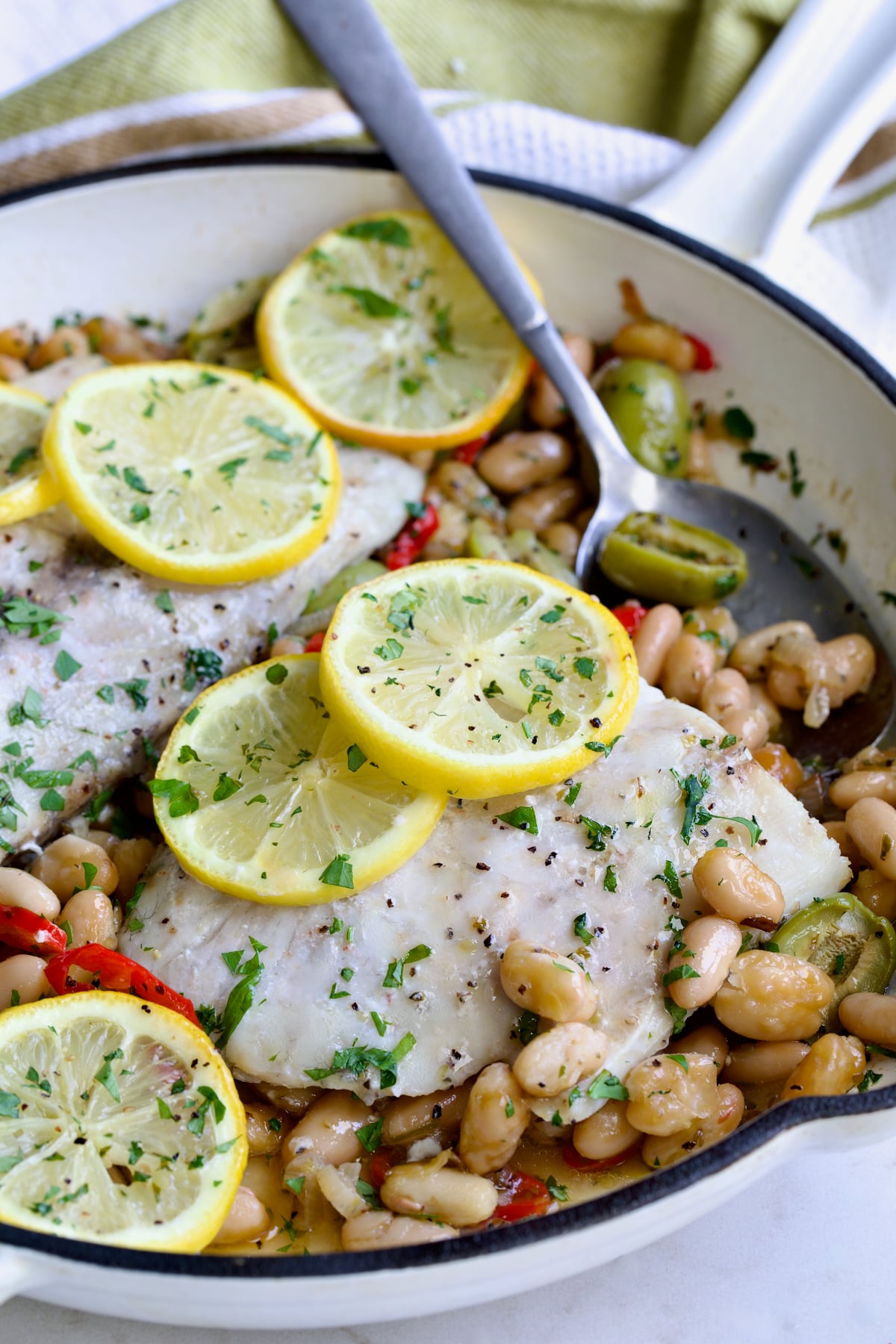 Mediterranean Fish and White Bean Skillet in white cast iron skillet topped with lemon slices with serving spoon.