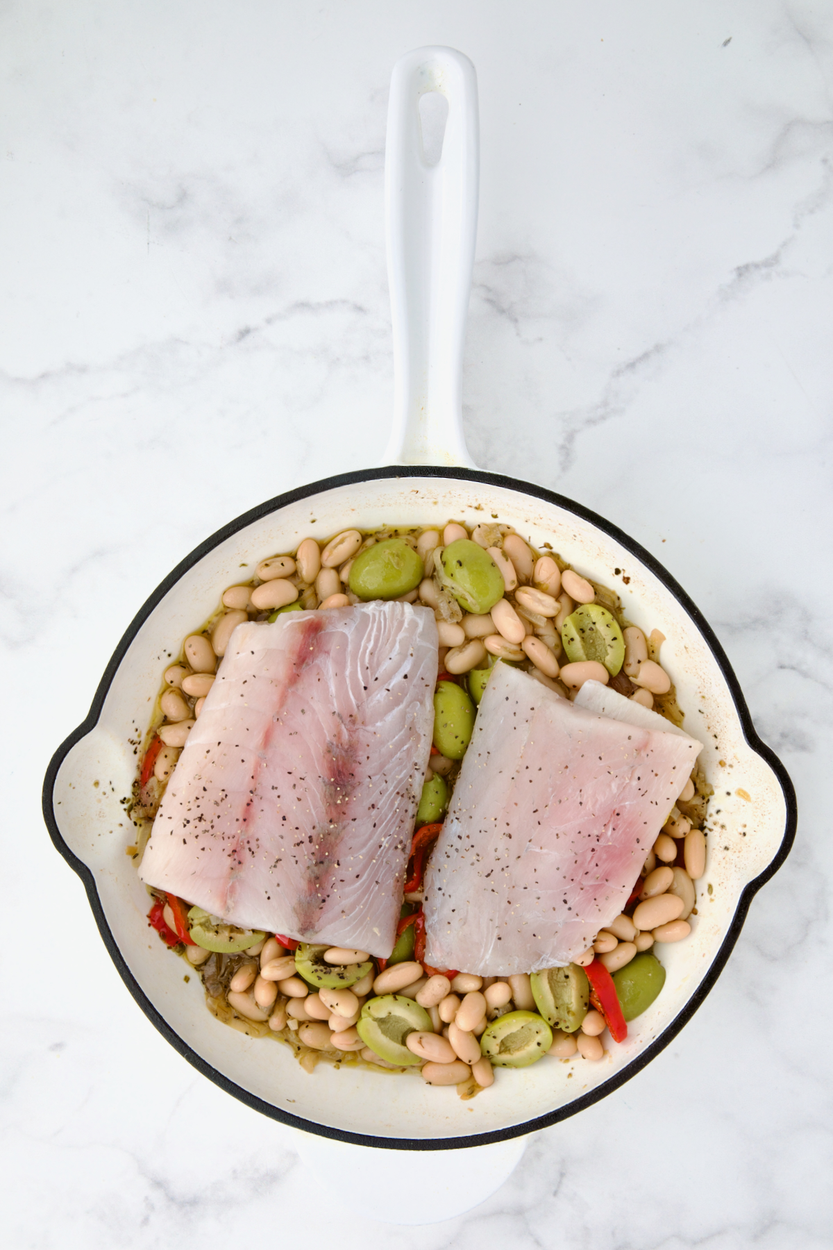 Uncooked fish fillets placed atop white bean mixture in white cast iron skillet.