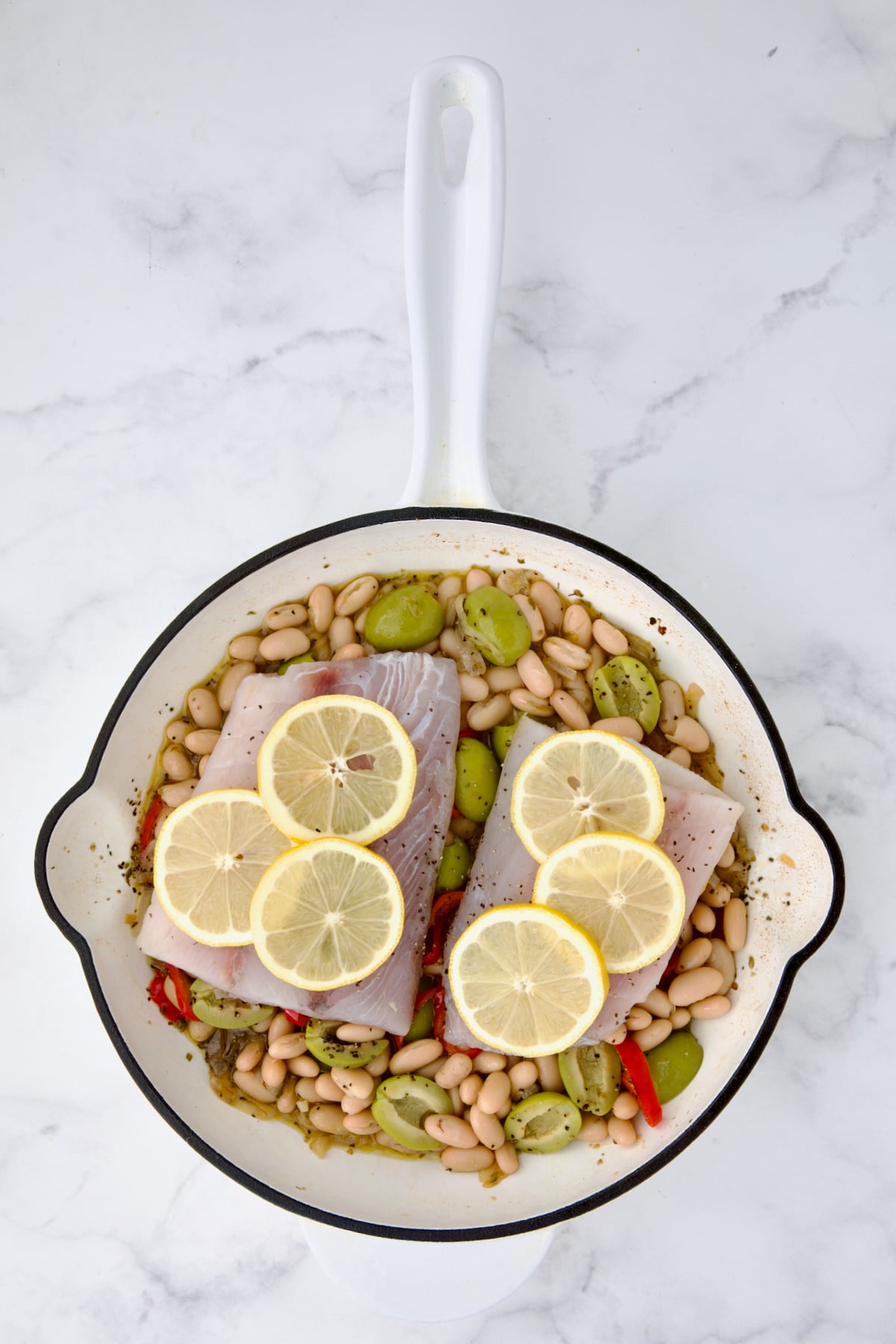 Uncooked fish fillets placed atop white bean mixture in white cast iron skillet with lemon slices added.